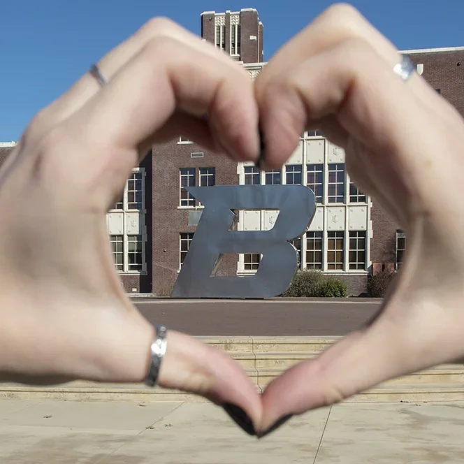 Hands form a heart shape framing a large metal letter "B" in front of a brick building.